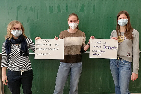 3 Schülerinnen stehen vor einer Tafel und halten 2 Schilder mit der Aufschrift. "weil Demokratie die Pressefreiheit schützt" und "weil wir unser Demokratie wertschätzen sollten"