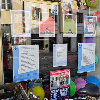 verschiedene Text und gemalte Plakate sind in einem Fenster zu sehen. Dazu bunte Luftballons und davor ein Fahrrad