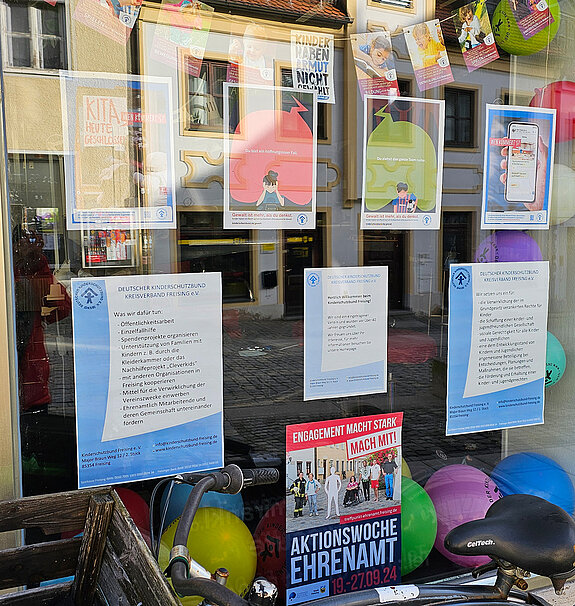 verschiedene Text und gemalte Plakate sind in einem Fenster zu sehen. Dazu bunte Luftballons und davor ein Fahrrad