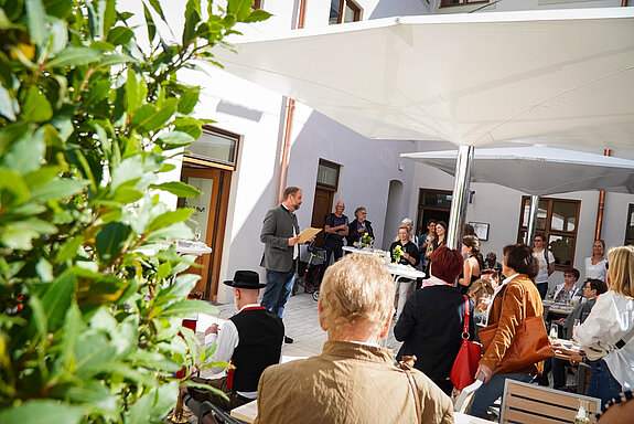 Auf dem Foto ist die Eröffnungsrede von Oberbürgermeister Tobias Eschenbacher vor dem dASAMa Dialograum zu sehen. Die Aufnahme wurde seitlich gemacht, um die Atmosphäre während der Rede einzufangen. Die Sonne scheint, und der Innenhof des dASAMa ist gut gefüllt. Die Stimmung wirkt angenehm und positiv.