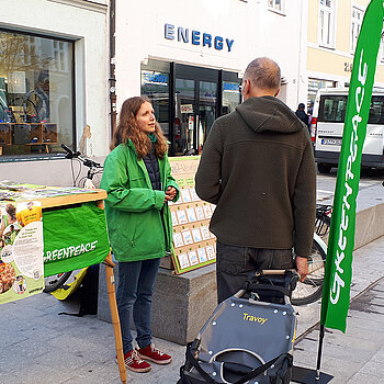 eine junge Frau zwischen zwei grünen Beachflags und in grüner Greenpeace-Jacke spricht mit einem Mann. 