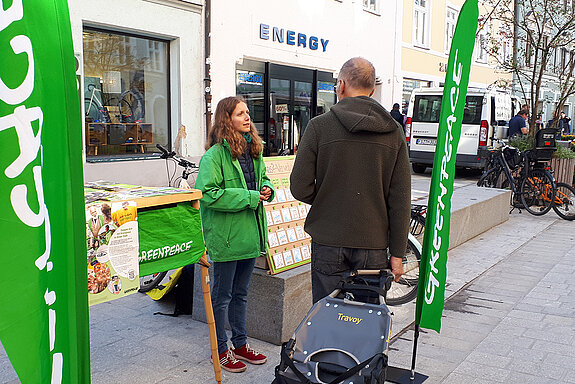 eine junge Frau zwischen zwei grünen Beachflags und in grüner Greenpeace-Jacke spricht mit einem Mann. 