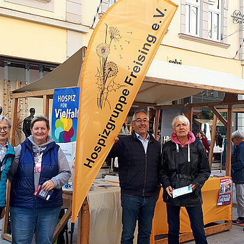 drei Frauen und ein Mann stehen vor einem Dultstand um eine orangefarbene Beachflag mit dem Logo: Hospizgruppe Freising