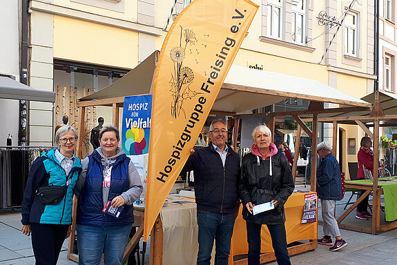 drei Frauen und ein Mann stehen vor einem Dultstand um eine orangefarbene Beachflag mit dem Logo: Hospizgruppe Freising