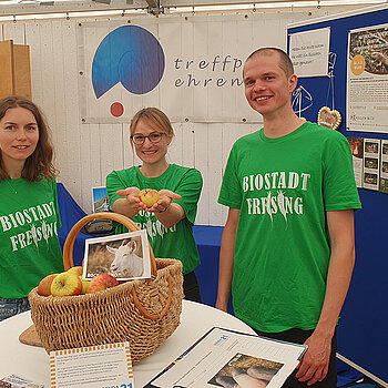 zwei Frauen und ein Mann in grünen T-Shirts mit Aufdruck "Biostadt Freising" stehen hinter einem Stehtisch mit Apfekorb darauf. Daneben eine Pinnwand mit Infos.