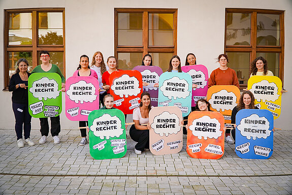 Auf dem Foto ist eine Gruppe von 14 Studierenden der Fachakademie für Sozialpädagogik Freising zu sehen. Sie nehmen an einer Aktion zum Weltkindertag teil. Die Studierenden halten Plakate in den Händen, auf denen Kinderrechte dargestellt sind. 