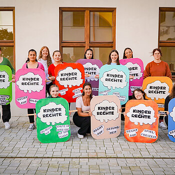 Auf dem Foto ist eine Gruppe von 14 Studierenden der Fachakademie für Sozialpädagogik Freising zu sehen. Sie nehmen an einer Aktion zum Weltkindertag teil. Die Studierenden halten Plakate in den Händen, auf denen Kinderrechte dargestellt sind. 