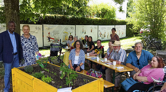 Ausstellungseröffnung mit Samuel Fosso und Manuela Wegler. Foto: Stadt Freising Eine Gruppe Menschen an Biertischen unter Bäumen, im Hintergrund Bauzaunbanner mit Cartoons, im Vordergrund ein Hochbeet.