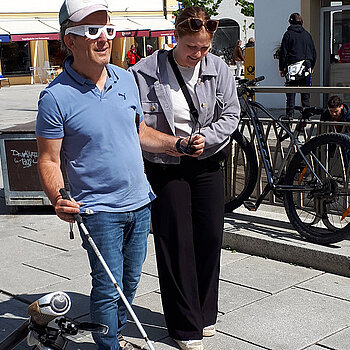 Ein Mann probiert das Laufen mithilfe der Leitlinie und einem Blindenstock aus. Foto: Stadt Freising Eine Frau hilft einen Mann mit Blindensimulationsbrille und Blindenstock