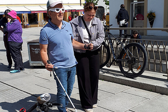 Ein Mann probiert das Laufen mithilfe der Leitlinie und einem Blindenstock aus. Foto: Stadt Freising Eine Frau hilft einen Mann mit Blindensimulationsbrille und Blindenstock