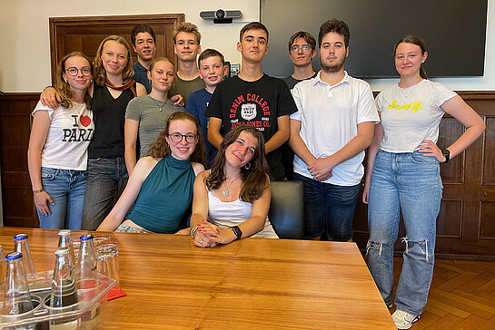 12 junge Leute in einem Sitzungsraum, 10 stehend, vorne am Tisch sitzend zwei junge Frauen.