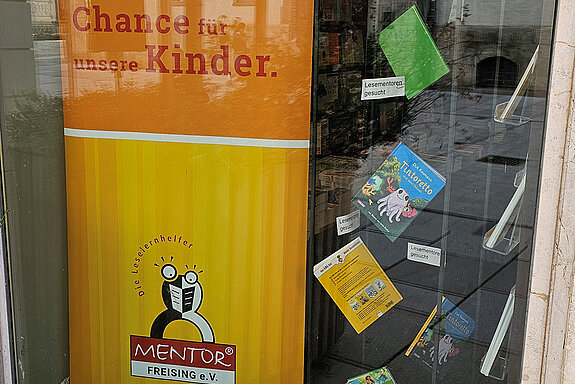 ein orange-gelber Aufsteller und daneben Kinderbücher wie ein Mobile aufgehängt. Dazwischen der Text "Lesementor gesucht"