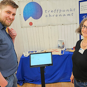 Ein Mann und eine Frau stehen neben einem kleinen Bildschirm, auf dem eine Frage angezeigt wird. Im Hintergrund das blau-rote Logo des Treffpunkts Ehrenamt