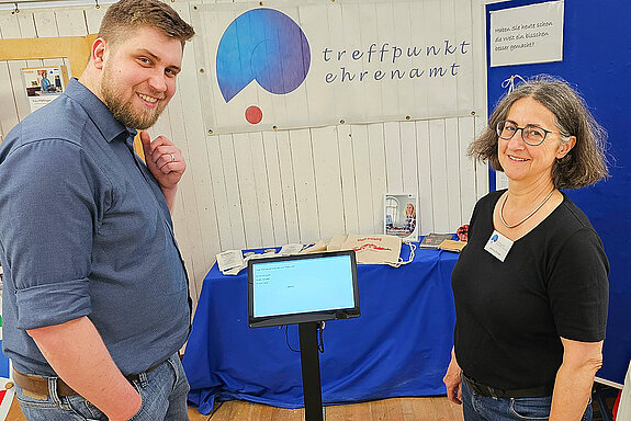 Ein Mann und eine Frau stehen neben einem kleinen Bildschirm, auf dem eine Frage angezeigt wird. Im Hintergrund das blau-rote Logo des Treffpunkts Ehrenamt