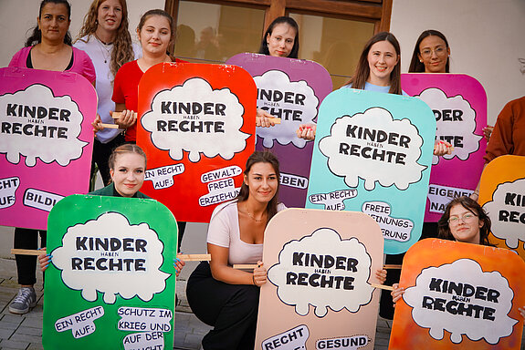 Auf dem Foto ist eine Gruppe von 10 Studierenden der Fachakademie für Sozialpädagogik Freising zu sehen. Sie nehmen an einer Aktion zum Weltkindertag teil. Die Studierenden halten Plakate in den Händen, auf denen Kinderrechte dargestellt sind. 