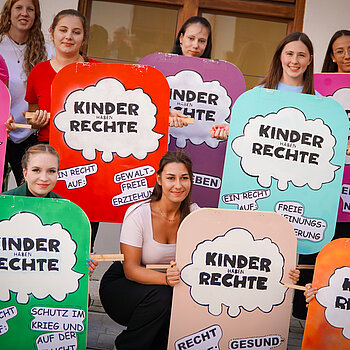 Auf dem Foto ist eine Gruppe von 10 Studierenden der Fachakademie für Sozialpädagogik Freising zu sehen. Sie nehmen an einer Aktion zum Weltkindertag teil. Die Studierenden halten Plakate in den Händen, auf denen Kinderrechte dargestellt sind. 