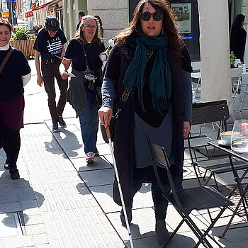 Stühle und Tische in den Leitlinien oder direkt daneben stellen ein Unfallrisiko dar. Foto: Stadt Freising Eine Frau mit Sonnenbrille und Langstock stößt auf einen Stuhl