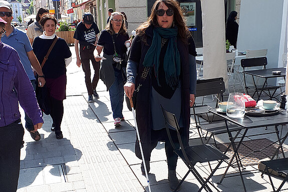Stühle und Tische in den Leitlinien oder direkt daneben stellen ein Unfallrisiko dar. Foto: Stadt Freising Eine Frau mit Sonnenbrille und Langstock stößt auf einen Stuhl