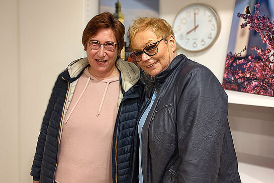 zwei Frauen mit Brille, freundlich lächelnd. Im Hintergrund eine Uhr und ein buntes Foto