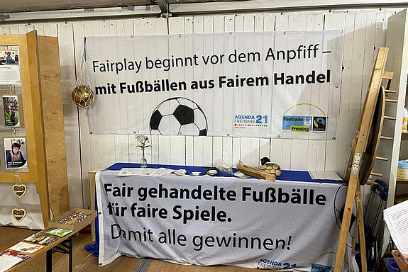 weiße Holzwand und Tisch mit Bannern mit Fußball und Fairtrade-Logo. "Fairplay beginnt vor dem Anpfiff - mit Fußbällen aus Rairem Handel. Fair gehandelte Fußbälle für faire Spiele. Damit alle gewinnen!" 