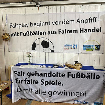 weiße Holzwand und Tisch mit Bannern mit Fußball und Fairtrade-Logo. "Fairplay beginnt vor dem Anpfiff - mit Fußbällen aus Rairem Handel. Fair gehandelte Fußbälle für faire Spiele. Damit alle gewinnen!" 