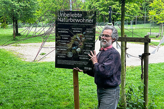 Ein Mann steht in einer Grünanlage und hält lächelnd ein großes Schild an einen Pfahl