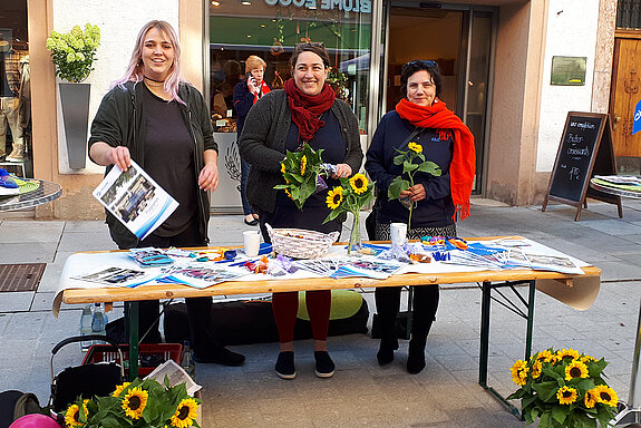 drei Frauen hinter einem Infotisch mit verschiedenen Materialien und geschmückt mit Sonnenblumen