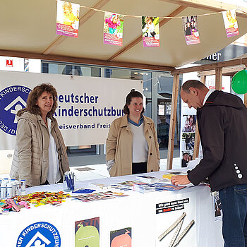 zwei Frauen stehen in einem Infostand mit Luftballons geschmückt. Ein Mann beugt sich über das Infomaterial