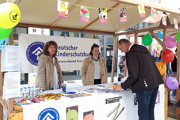 zwei Frauen stehen in einem Infostand mit Luftballons geschmückt. Ein Mann beugt sich über das Infomaterial