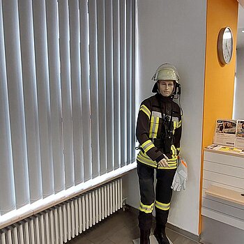 Eine lebensgroße Puppe in Feuerwehrmontur in einer Ecke stehend.