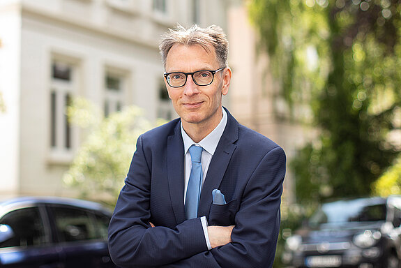 Ein Mann mit Brille im blauen Anzug mit hellblauer Krawatte und Brille. Im Hintergrund ein Stadthaus, Auto, Straßenbäume