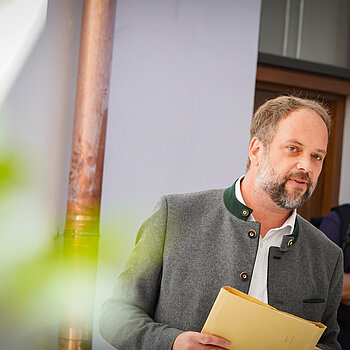 Auf dem Foto ist Oberbürgermeister Tobias Eschenbacher zu sehen. Es ist sonniges Wetter. Er hält einen dünnen Papierordner in der Hand. Im Hintergrund steht eine weitere Person. Tobias Eschenbacher trägt passend zum Anlass einen Trachtenjanker.