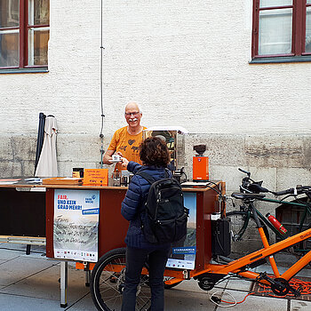 Ein lachender Mann reicht einer Frau ein Espresso-Tässchen über einer Theke, die gleichzeitig ein Fahrrad-Anhänger ist.