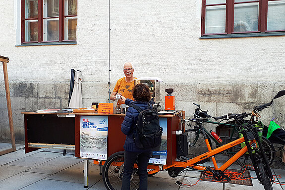 Ein lachender Mann reicht einer Frau ein Espresso-Tässchen über einer Theke, die gleichzeitig ein Fahrrad-Anhänger ist.